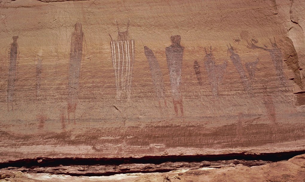 A row of tall Barrier Canyon Style figures, one with white vertical stripes, along a sandstone wall above a ledge
