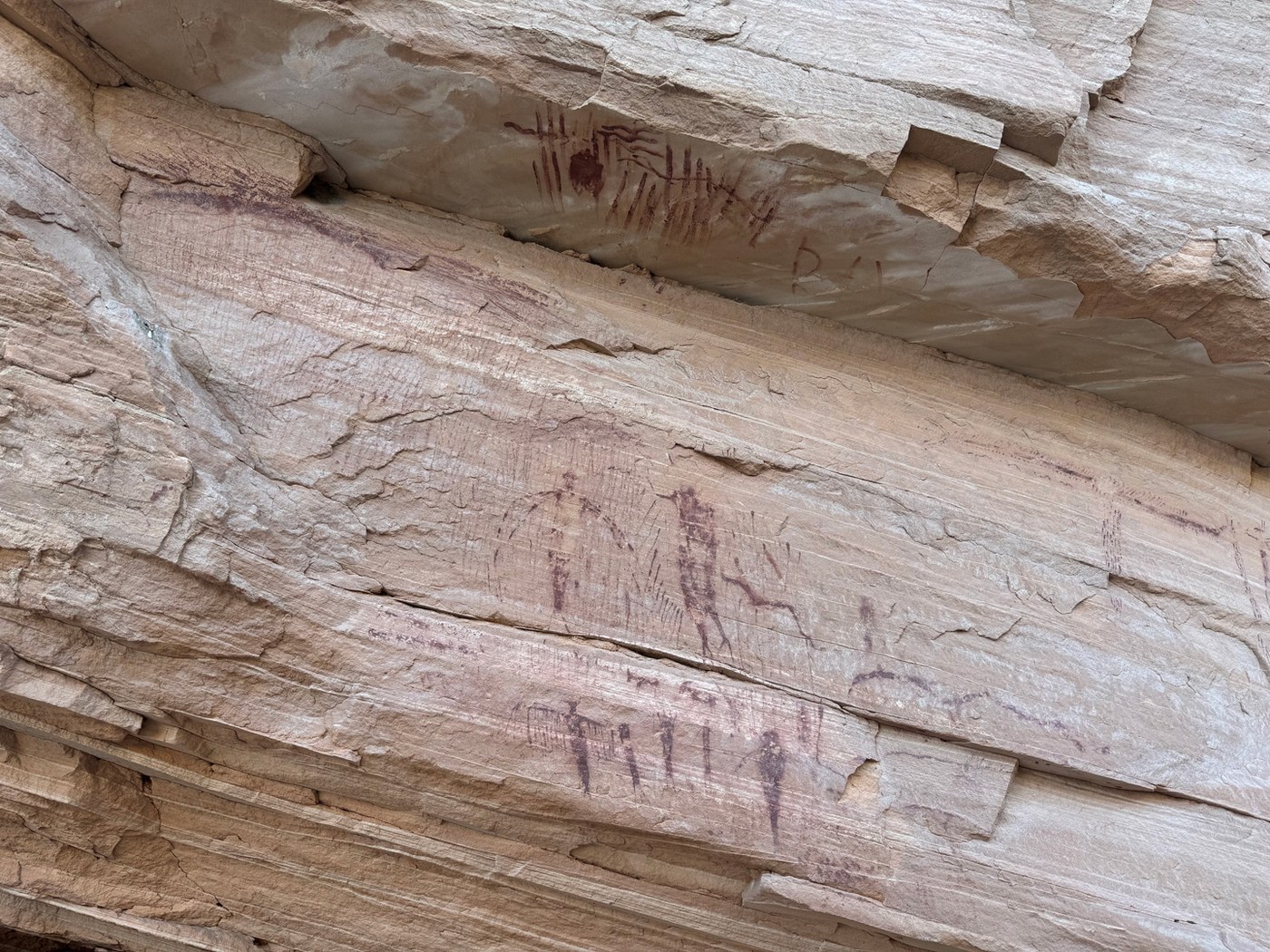 Small faded red figures and striped marks on ledges of layered sandstone below an overhang