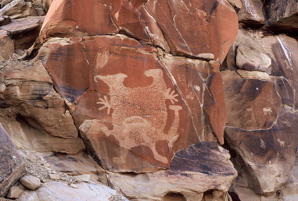 Large pecked Interior Line Style figure on a red rock face, with arms or branches at its sides and small figures on the rocks at right